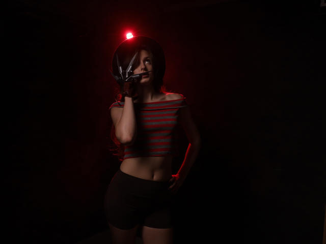 JasmineNeal with a red light on her helmet, holding a gun to her mouth, wearing a striped red and black crop top and black shorts, in a dark setting.