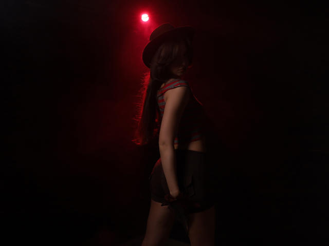 JasmineNeal with dark hair in a ponytail, wearing a hat and red-striped top, has her hand on her black skirt, standing in dim red light, looking back.