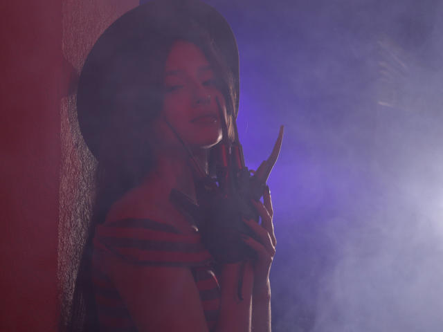JasmineNeal with a huge black hat, holding Freddy Krueger's hand with sharp claws, wearing a red and black striped shirt, in a smoky purple and red light.