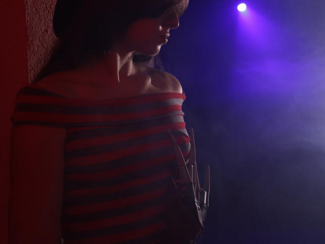 JasmineNeal with dark hair, off-shoulder red and gray striped shirt, light from purple-blue spotlight, shadow on right side, holding a black phone, red wall on left.