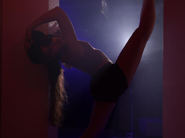 JasmineNeal with long brown hair, tattoos on her side, arms raised, wearing black shorts, legs in a split, backlit by blue and pink lights, standing in a dark room.