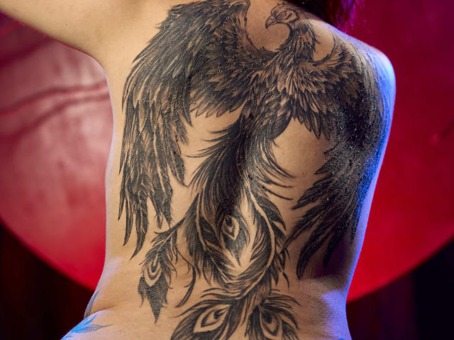 Le tatouage sur le dos de Cataleya Sweet représente un phénix noir détaillé, avec des plumes fluides et un motif d'œil de paon, rehaussé d'un éclairage bleu et rouge..