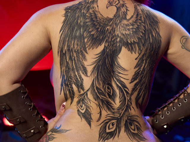 Le dos de Cataleya Sweet est orné d'un grand tatouage de paon très détaillé, avec des plumes qui descendent le long de sa colonne vertébrale. Elle porte des bottes marron à lacets et a les mains sur ses fesses..