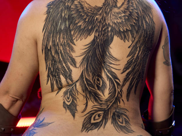 Le tatouage sur le dos de Cataleya Sweet représente de grandes plumes noires avec des yeux de paon, recouvrant tout son dos supérieur. Elle porte des manchettes en cuir marron. Le tatouage est détaillé et foncé..