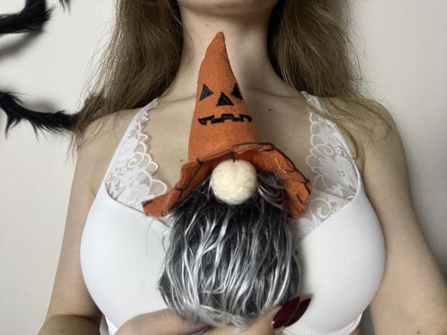 MiraRosse's tits are covered by a white lace bralette, wearing a pumpkin gnome hat with black hair and white pom-pom, in a selfie.