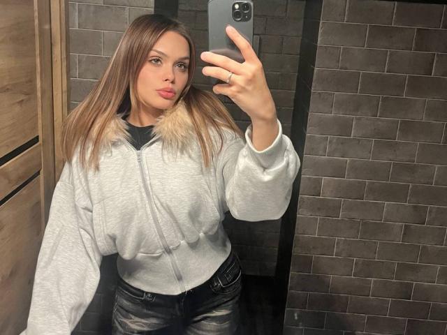 DelphaHerzfeld, com cabelos castanhos longos, lábios carnudos, vestindo um moletom cinza com detalhes de pele sintética e calças jeans pretas, tirando uma selfie no espelho com a mão direita..