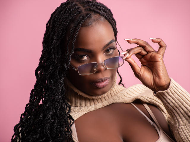 Nyla Parker, con la pelle scura, lunghi capelli trecciati, che indossa occhiali, un maglione beige, che mostra il décolleté, sfondo rosa, che tiene occhiali con unghie curate. Seno visibile, espressione concentrata, aspetto elegante..