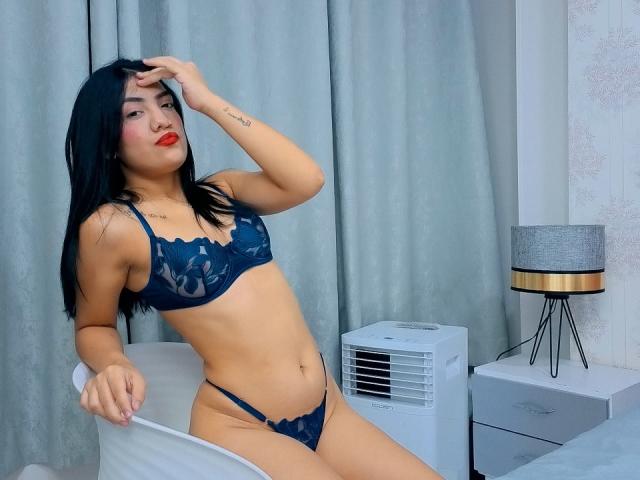 CamilaFroes with black hair, wearing dark blue lace lingerie, sitting on a chair, hand on head, red lipstick, small tattoos on arm, white fan and grey lamp in background.