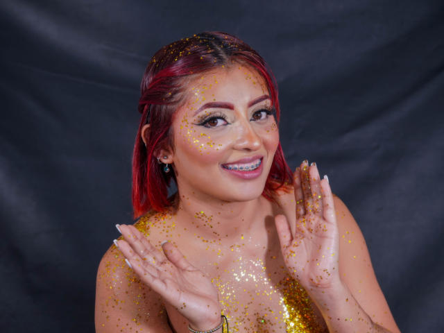 LizBonnet with red hair, glitter on face and hands, smiling, hands up, glittery top, dark background, visible tits, glitter on chest, earrings, pink lipstick, digital photograph.