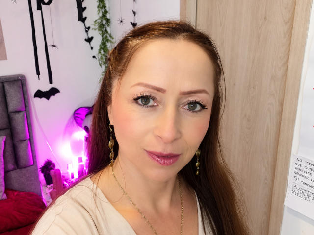 GianineLarose tiene la piel clara, ojos verdes y cabello largo castaño. Lleva lápiz labial rosa y aretes dorados, con un collar dorado. Sus senos son pequeños y está sonriendo..
