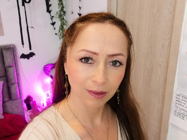 GianineLarose tiene piel clara, cabello castaño, ojos verdes y lápiz labial rosa. Lleva una blusa blanca, aretes dorados y un collar. Sus pechos son pequeños. Está haciendo una cara linda..