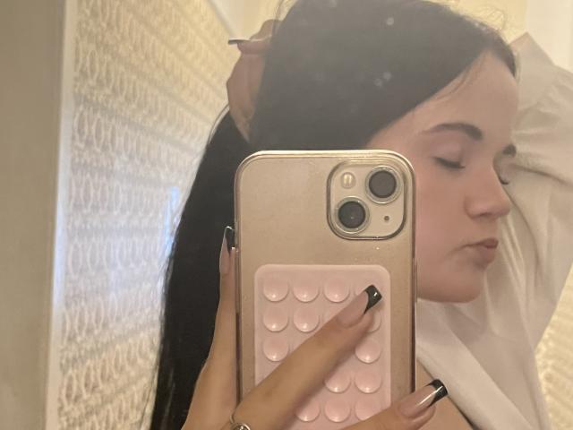 RoxyVesper with long black hair, holding a gold iPhone with pink grip, taking a mirror selfie, eyes closed, hand in hair, wearing white shirt, nails painted black.