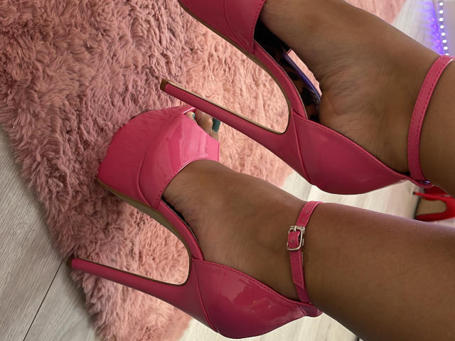 MoniqueBliss's shiny pink high heels with ankle straps, standing on a pink fluffy rug, showing her dark brown, smooth, and well-groomed feet.