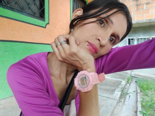 TiffannyConors with pink lipstick, black hair in a ponytail, purple top, pink watch, and lip piercing, resting chin on hand, showing ring and wrist. Boob outline visible.
