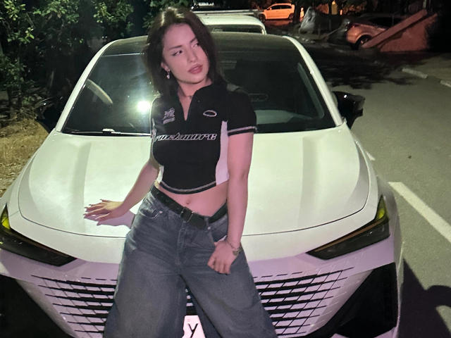CharlotteDegrande, avec une poitrine de taille moyenne, un crop top noir et un jean bleu, appuyée contre une voiture blanche la nuit, regardant légèrement sur le côté avec une expression détendue..
