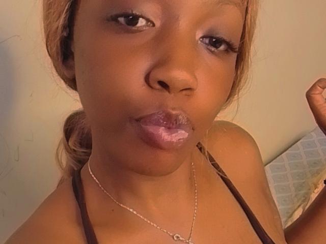SweetPunani has dark brown skin, pouty pinkish-purple lips with white lipstick, and is wearing a brown strap top. She has a thin necklace and a slight smile.