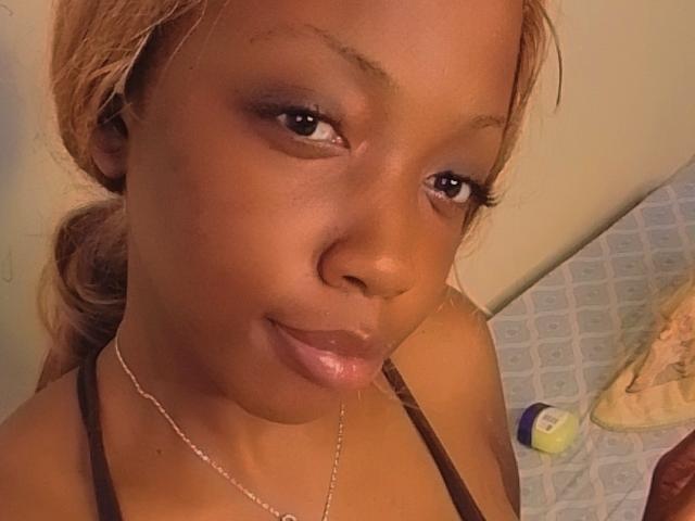 A selfie of SweetPunani with dark brown skin, glossy pink lips, and brown eyes. She's wearing a brown top and a silver necklace, with her hair pulled back.