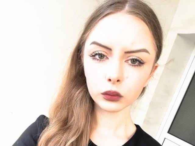 LourdesWhilby with a serious face, light brown hair in a ponytail, dark red lipstick, and thick eyebrows. She's wearing a black top, looking straight into the camera.