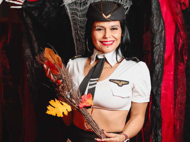 CharlotteCrofort, with black hair and red lipstick, wearing a sexy pilot uniform, holding autumn leaves, smiling, has large tits, and is standing in front of dark red and black drapes.