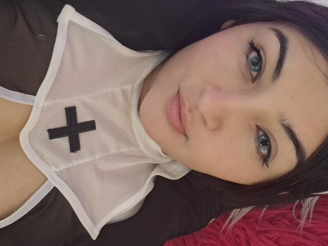 LindaSophieX, con ojos azules, cejas negras y labios rosados, vestida con un conjunto transparente negro y blanco con una cruz en el pecho, acostada sobre una superficie roja..