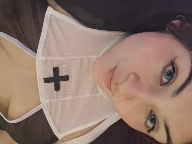 LindaSophieX with blue eyes, pink cheeks, and black cross on white mesh top, showing cleavage, wearing black and white outfit, lying down, looking at camera with slight smile.