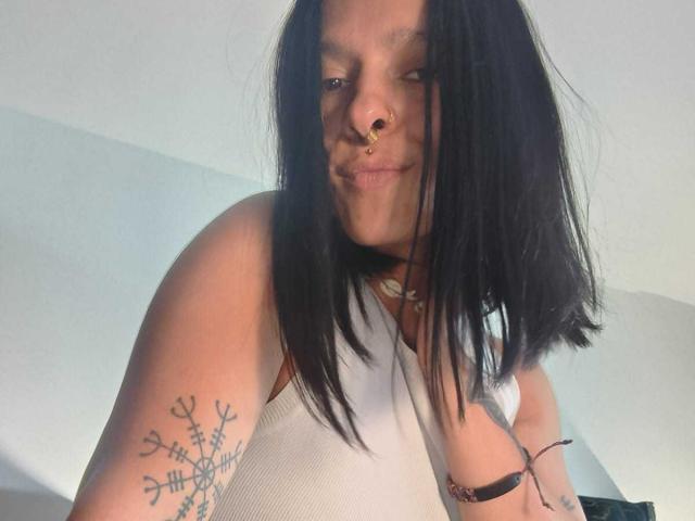Amelie Blake, con lunghi capelli neri, un piercing al naso, una canotta bianca che mostra un tatuaggio sulla parte superiore del braccio, sorridente, con la pelle chiara, un trucco leggero e una spallina del reggiseno nero visibile sul lato destro..