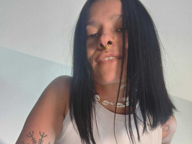 Amelie Blake, con capelli neri, piercing al naso, che sorride, indossa una canotta bianca, una collana, ha tatuaggi sulle braccia, ha la pelle chiara, guarda in basso, è un selfie, ripresa dalla parte superiore del corpo, con il seno visibile..