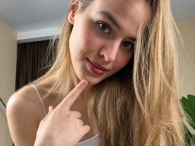 SandiWimbs with blonde hair, fair skin, and pink lips, holding her finger to her chin, smiling softly, wearing a white spaghetti-strap top, in a casual selfie.