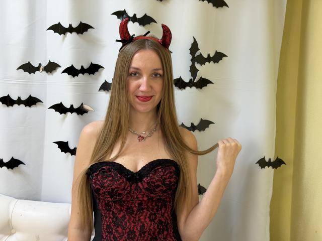 EllaMint with long brown hair, red and black lace corset, small red devil horns, holding her hair, smiling, wearing a red heart necklace, standing in front of a white curtain.