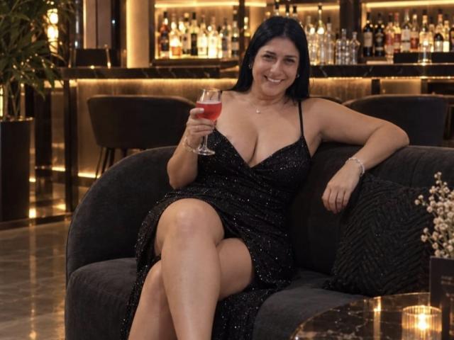 DamaraFoxy with big tits, black sparkly dress, holding red drink, sitting on dark couch, smiling, black hair, tan skin, bar background, crossed legs, cocktail glass in hand.