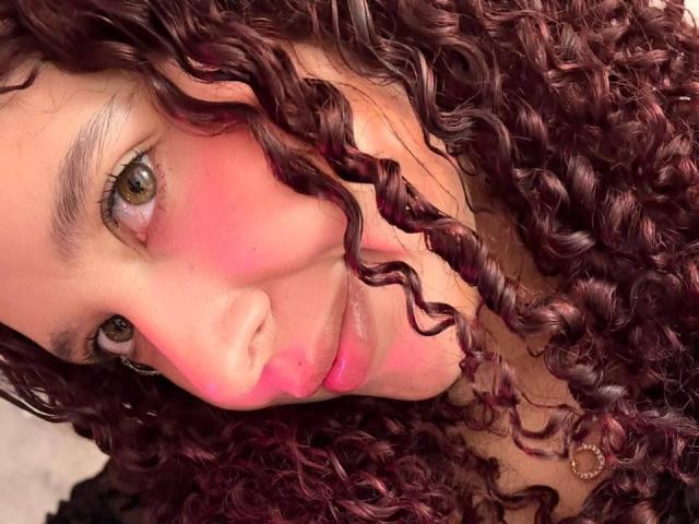 ValeryScot's face with curly, dark purple hair, pink blush on cheeks, brown eyes, and a slight smile. Her lips are slightly parted, showing a hint of pink.