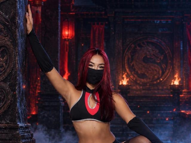 SashaHans with red hair, black mask, sports bra, and arm sleeves, arms up, holding column, showing toned abs, fiery background. Her tits are small, face is sexy.