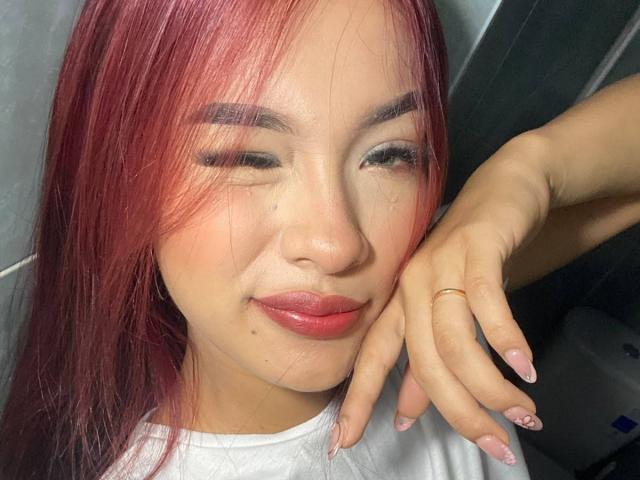 SashaHans with red hair, closed eyes, and pink lipstick, smiling slightly. She's wearing a white shirt, and has a gold ring on her right hand with pink nails.