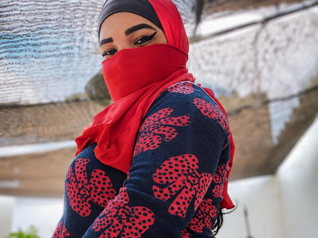 FalakYasin with a red hijab and mask, wearing a navy blue sweater with red polka dot bows, looking over her shoulder, showing her ass, with bold eyeliner.