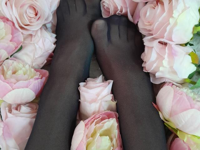 AmeliaMaiv's black pantyhose-covered feet nestled among pink and white roses. Soft petals and delicate flowers surround her toes. Sheer texture of the pantyhose contrasts with rose petals.