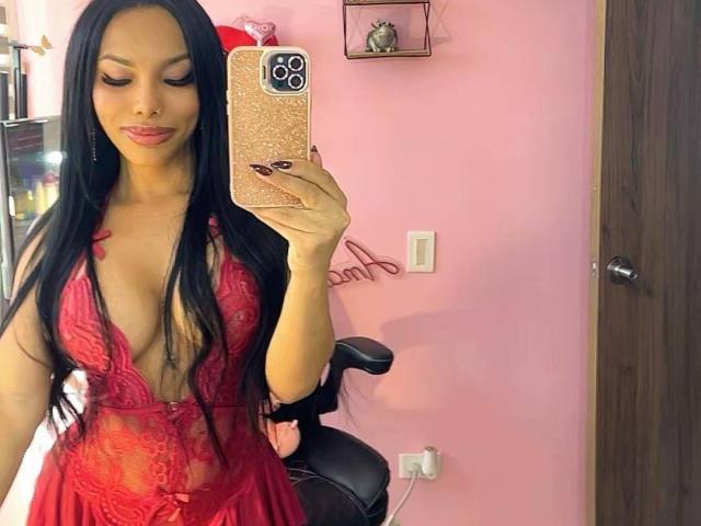 Amaranta69s with long black hair, red lace lingerie, and cleavage, holding a gold phone, taking a mirror selfie, showing her tits and red lipstick.