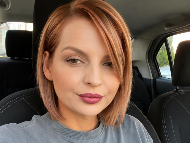 DouceAmelia has short, dirty blonde hair, pink lipstick, and a grey shirt in a car, looking slightly to the side with a subtle smile. Boobs not visible.