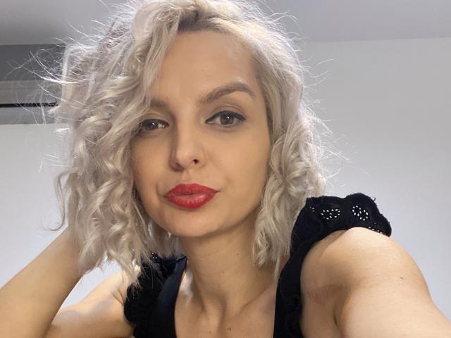 DouceAmelia with curly white hair, red lipstick, and black lace top, looking at the camera with a slight smile, hands in her hair. Her tits are small and she's cute.