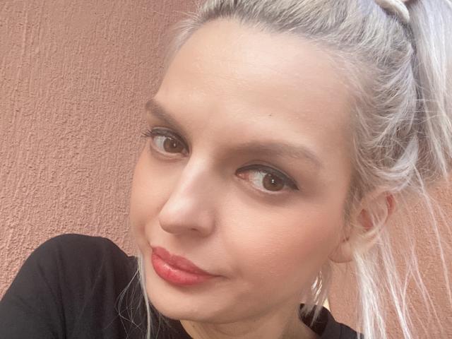 DouceAmelia has light blonde hair in a ponytail, brown eyes, red lipstick, and a slight smile. She's wearing a black shirt and gold earrings. Her skin is fair.