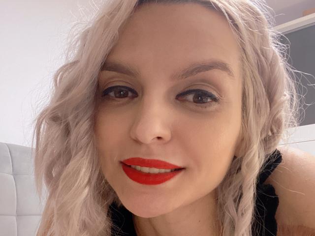 DouceAmelia with wavy, platinum blonde hair, red lipstick, and black tank top, smiling at the camera, showing her nose and eyes, with light makeup and subtle eyeliner.