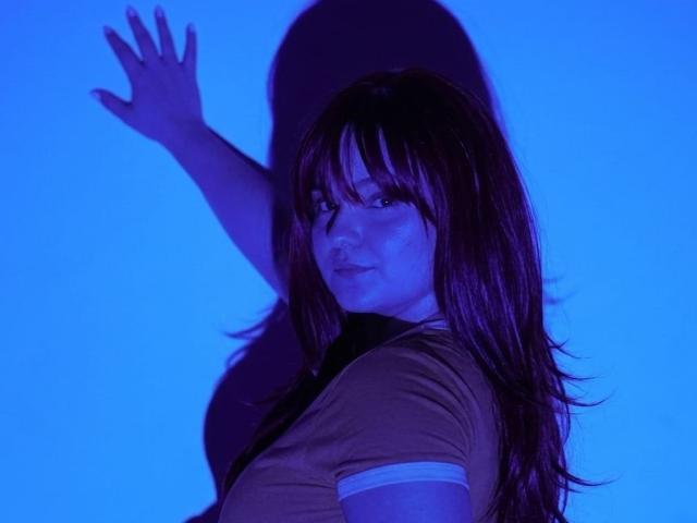 Maddy Jones, met lang bruin haar, die een gele blouse draagt, haar hand omhoogstekend, een schaduw werpend, onder blauw licht, kijkt terug met een licht geïrriteerde uitdrukking..