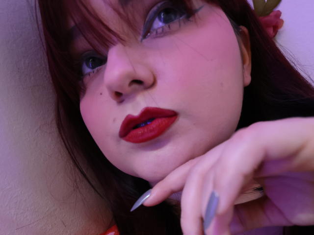 Maddy Jones tiene una piel rosada, lápiz labial rojo y uñas largas y oscuras. Su rostro está cerca de la cámara, mostrando su nariz y lunares, con su mano en primer plano..