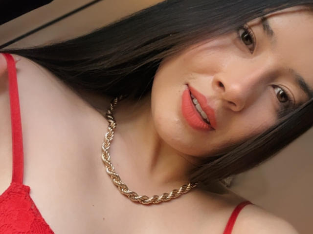 Emma Lombardi a de longs cheveux noirs, du rouge à lèvres, et porte un soutien-gorge en dentelle rouge avec un collier en chaîne dorée. Ses seins sont visibles. Elle sourit légèrement..