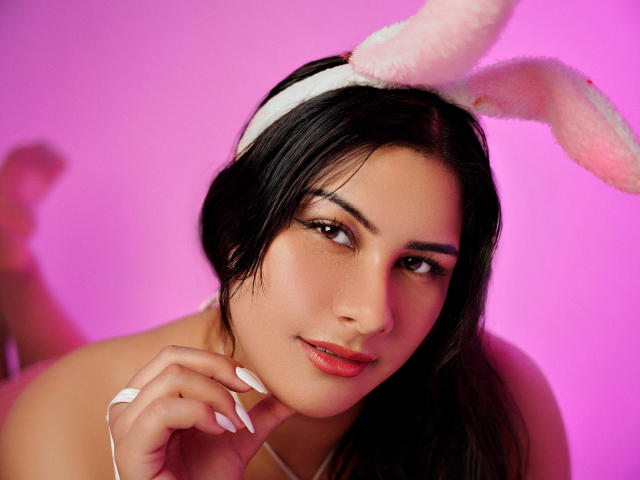 BonnieGray with dark hair, pink bunny ears, and white headband, smiling with pink lips, holding her chin, pink background, long white nails, sexy, cute, soft lighting.