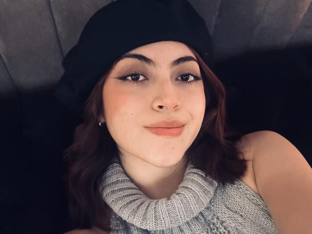 SpencerLe has a cute face, brown wavy hair, black beanie, and wears a grey knitted turtleneck, smiling with subtle pink lipstick, looking up with black eyeliner.