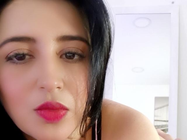 NicollJeans has dark black hair, brown eyes, pink lipstick, and a slight pout. She's wearing a pink top, with her bare shoulder visible. Her face is centered in the photo.