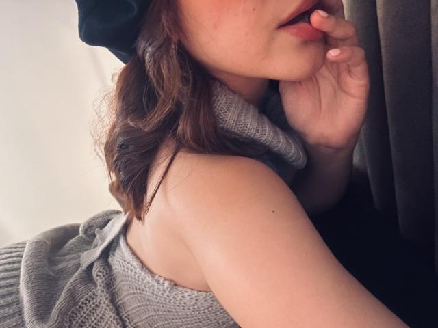 SpencerLe with wavy brown hair, wearing a green beanie, gray knitted sweater, biting her finger, showing her bare shoulder and upper chest. Her hand is near her mouth.