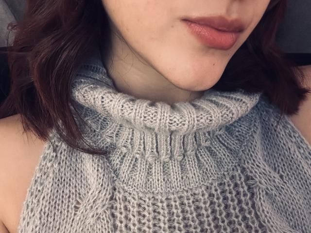 SpencerLe's face with pinkish lips, wearing a grey, knitted turtleneck sweater. Her brown wavy hair is visible. The sweater's texture is thick and patterned.