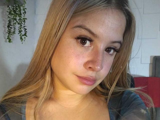 MelaniBecker has blonde hair, brown eyes, and a cute face with light freckles. She's looking at the camera with a subtle smile, wearing a blue top.