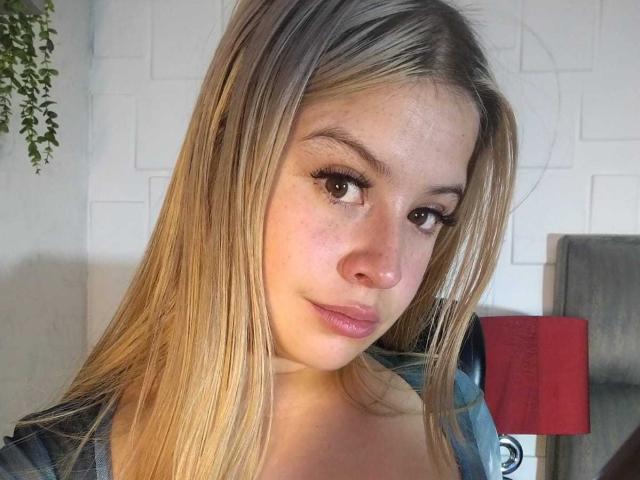 MelaniBecker with long blonde hair, brown eyes, and pink lips, wearing a blue shirt, looking at the camera with a slight smile, showing her cute nose and small tits.